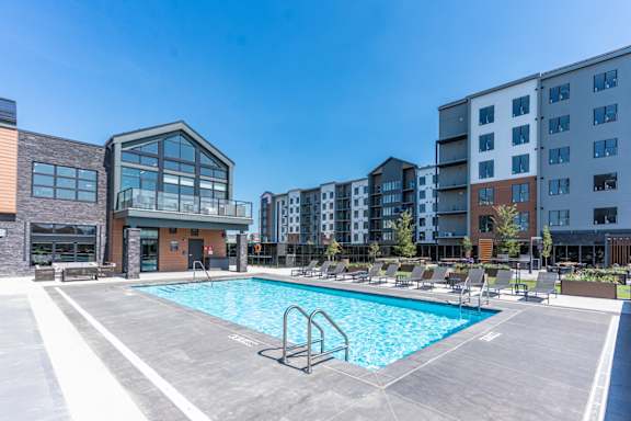 A swimming pool in a sunny day surrounded by chairs and buildings.