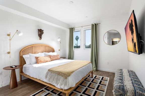 Bedroom with a bed, a side table, a rug, a television, lamps, and windows with curtains.