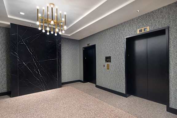 A hallway with a chandelier, marble wall, and two elevators.