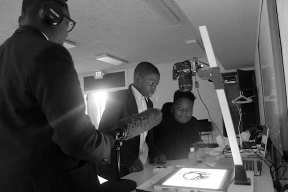 A man in a suit is holding a microphone while standing next to a young boy who is sitting at a desk with a computer.