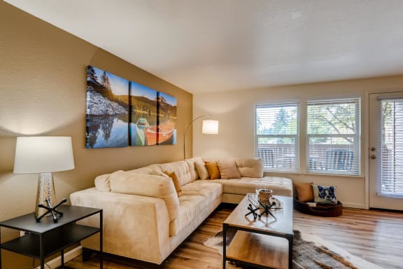 Apartments Near Happy Valley For Rent - Sunnyside Village - a Living Room with Wood-Style Flooring, a Large Plush Couch, a Coffee Table, a Small Table with a Lamp, Large Windows with White Blinds, and a Door to the Private Patio
