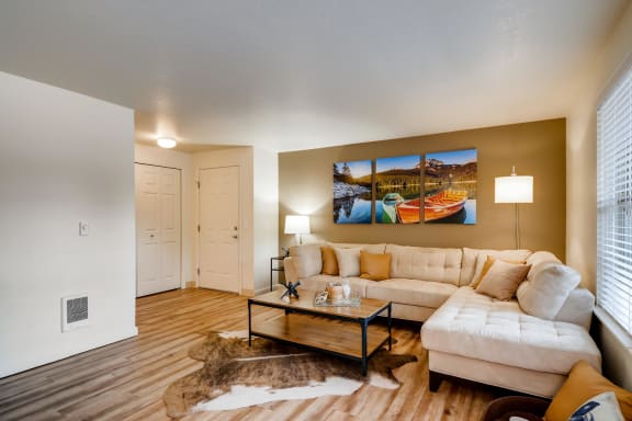 Apartments in Happy Valley, OR, for Rent - Sunnyside Village - Spacious Living Room with Wood-Style Flooring, Animal Skin Rug, Cream-Colored Sectional Couch, Wooden Coffee Table, Floor Lamp, Side Table with Lamp, White Walls with a Beige Accent Wall, and Large Window.