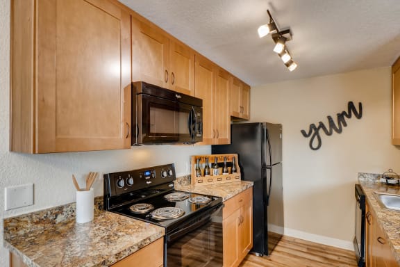 Apartments Happy Valley, OR - Sunnyside Village - a Kitchen with Wood-Style Flooring, Granite Countertops, Black Appliances Including a Refrigerator, Oven, Dishwasher, and Built-In Microwave, Brown Wood-Style Cabinetry, and an Overhead Lighting Fixture