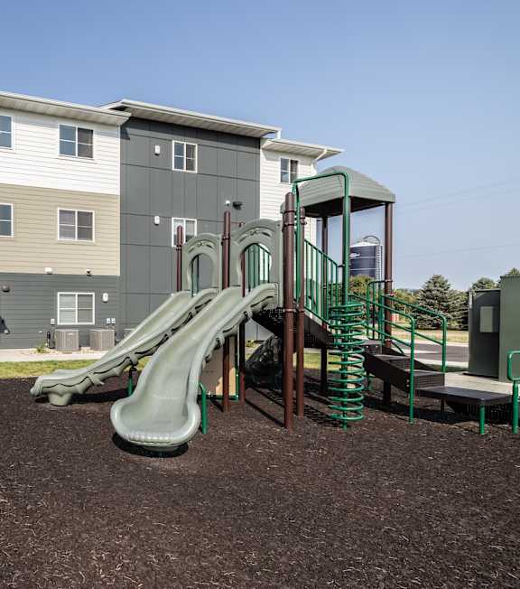 A playground with a green slide in front of a two-story apartment building.