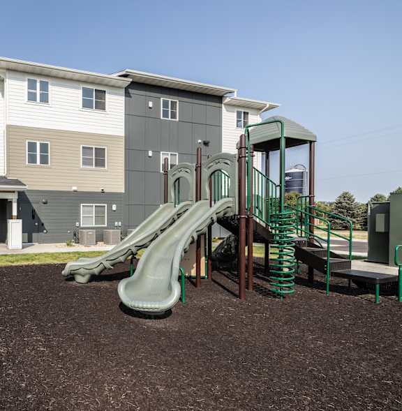 A playground with a green slide in front of a two-story apartment building.