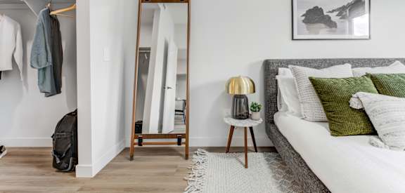 A bedroom with a bed, a clothes rack, a side table with a lamp, and a painting on the wall.