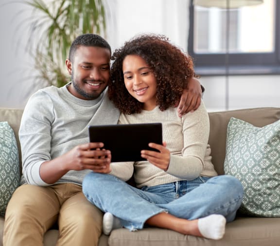 A couple is sitting on a couch, with the man holding a tablet and the woman sitting next to him.