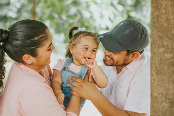 A family of three is standing outdoors with a tree in the background.