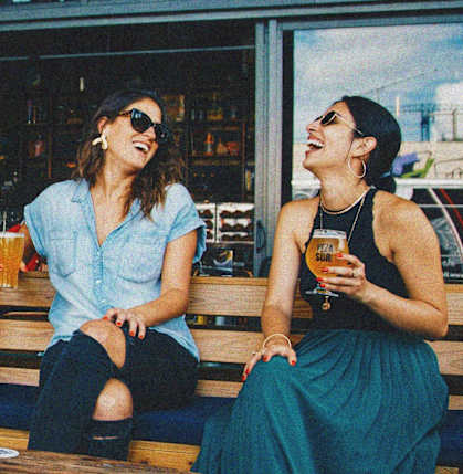 two women sitting on a bench laughing and drinking beer