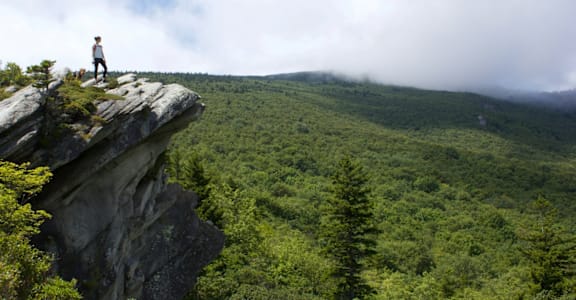 A person stands on a cliff edge overlooking a green valley.