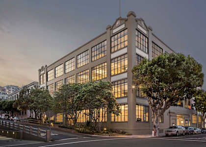 A large, modern building with a lot of windows is surrounded by trees and has a parking lot in front.