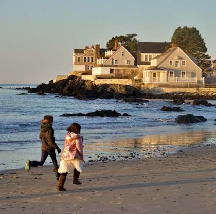 two girls running on the beach near the ocean with a house in the background