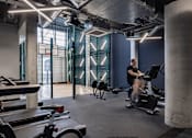 Thumbnail 6 of 8 - A man is working out on a stationary bike in a gym with a concrete column and a row of treadmills.