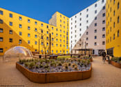 Thumbnail 3 of 8 - A modern building with yellow and white exterior and a courtyard with a tree and a dome-like structure.