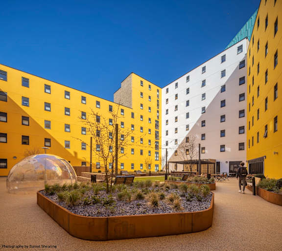 A modern building with yellow and white exterior and a courtyard with a tree and a dome-like structure.