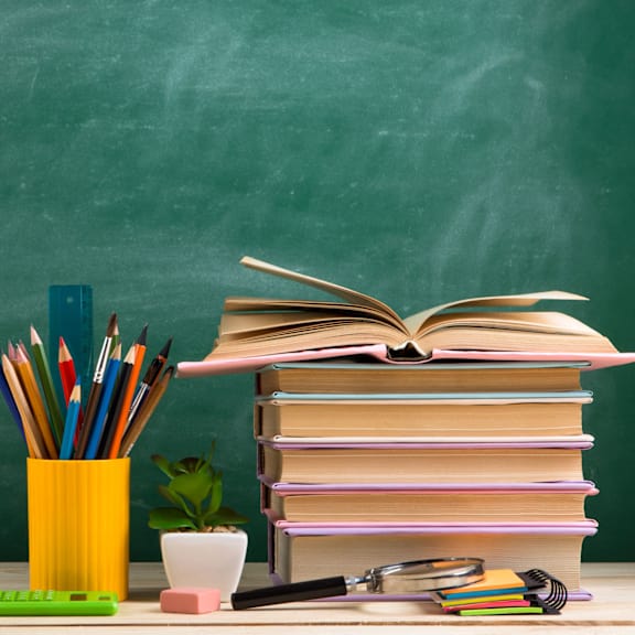 A stack of colorful books with a magnifying glass and pencils on a desk.