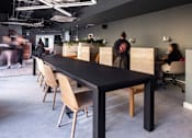 Thumbnail 8 of 8 - A long black table with tan chairs in a room with people walking in the background.