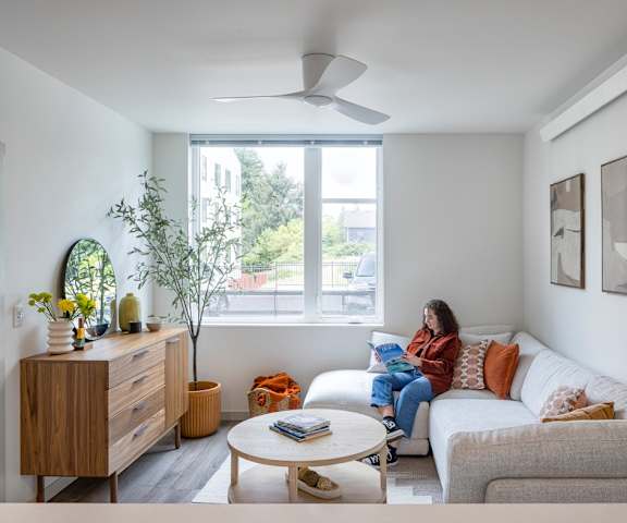 A woman is sitting on a couch in a living room with a bed, a table, and a plant.