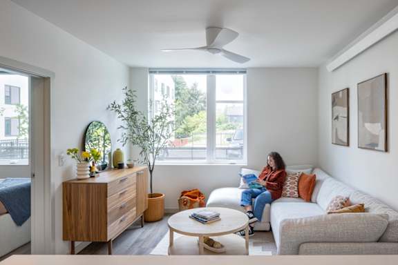 A woman is sitting on a couch in a living room with a bed, a table, and a window.