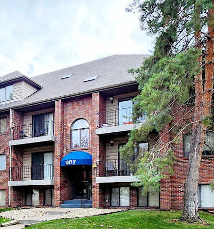 A three-story brick building with a blue awning and a tree with orange leaves in front.