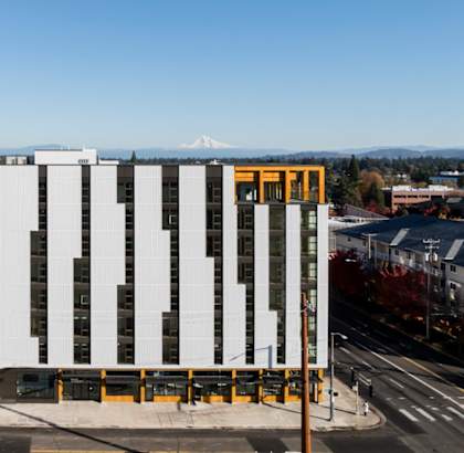 A modern building with a white facade and a yellow entrance is surrounded by other buildings and trees.