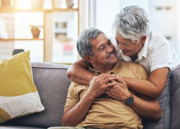 An older man and woman are sitting on a couch, with the woman leaning in to give the man a hug.