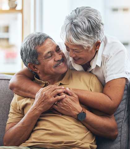 An older man and woman are sitting on a couch, with the woman leaning in to give the man a hug.