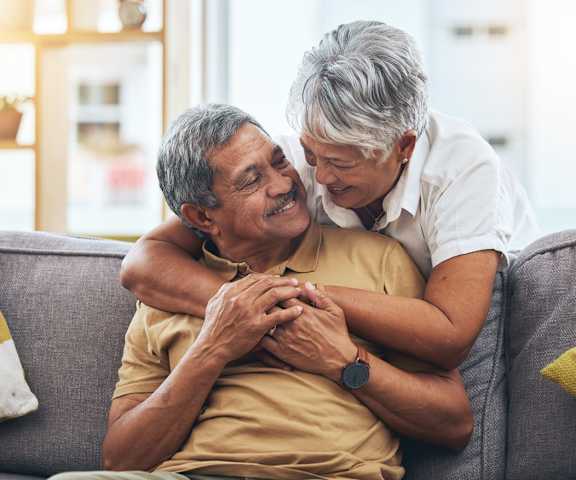 An older man and woman are sitting on a couch, with the woman leaning in to give the man a hug.