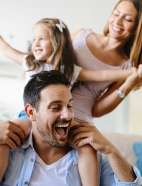 A man is sitting on a couch with a little girl on his shoulders and a woman standing behind them both.