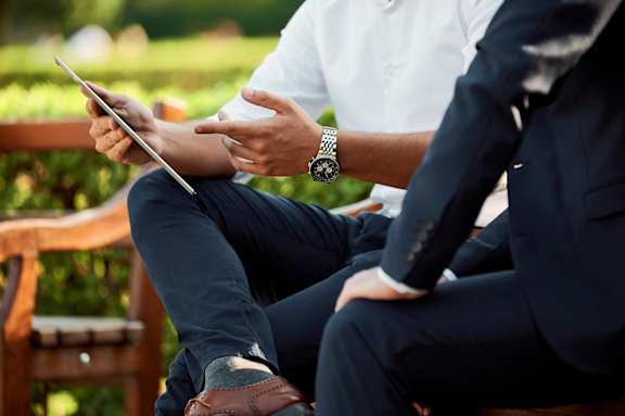 Two men in suits are sitting on a bench, one holding a tablet.