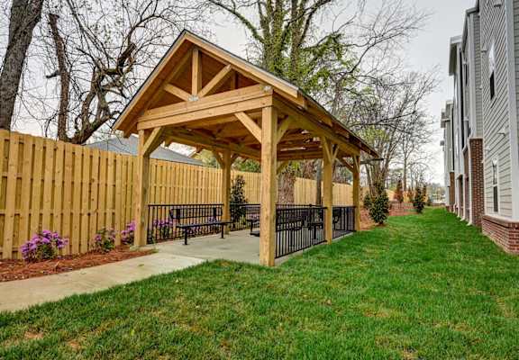 A wooden pavilion with a black fence and benches is surrounded by a grassy area.