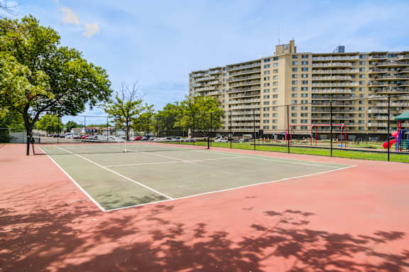 A tennis court with a red surface and white lines, surrounded by trees and apartment buildings at Premier at Cherry Hill, Cherry Hill, NJ