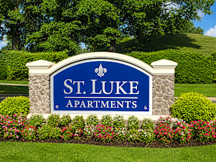 A sign for St. Luke Apartments is surrounded by flowers and greenery.