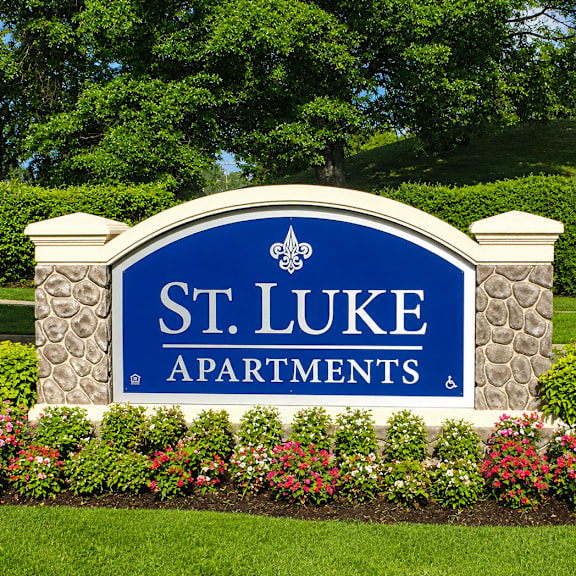 A sign for St. Luke Apartments is surrounded by flowers and greenery.
