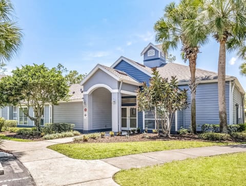 The exterior of a leasing office with trees, palm trees, a sidewalk, and grass areas.