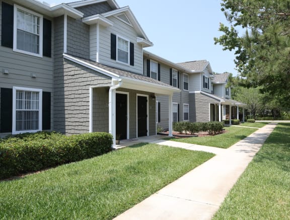 The resident building is a grey shingle sided building with white trimmed windows. In front of the entrance is a concrete walkway that extends to both sides to a sidewalk. The landscaping on either side of the entrance is lush green grassy plants, brown mulch, and large green bushes.