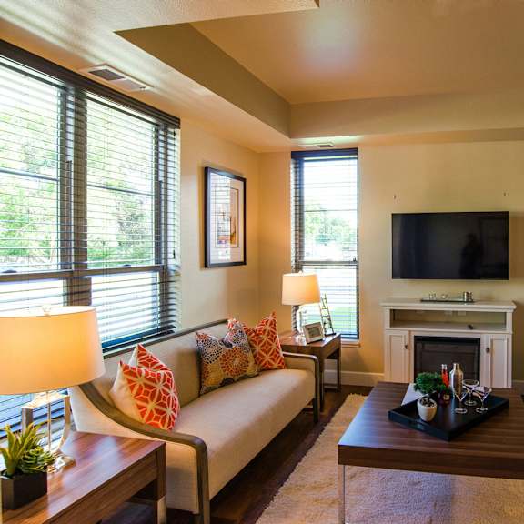 Siena Living Room with Fireplace The spacious living room with a couch, orange throw pillows, large windows, a ceiling fan, wall art, plush chair, coffee table, decorations, large windows, artwork, and a TV at Siena Apartment Homes.
