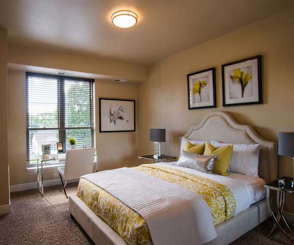 A spacious bedroom with a queen-size bed with a yellow and white bedspread, a dresser, wall art, lamps, decorations, two windows, and nightstands with lamps at Siena Apartment Homes. 
