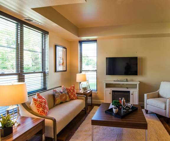 The spacious yellow living room with a tan couch, orange throw pillows, large windows, wall art, plush chair, coffee table, a cozy fireplace, decorations, large windows, and a TV at Siena Apartment Homes. 
