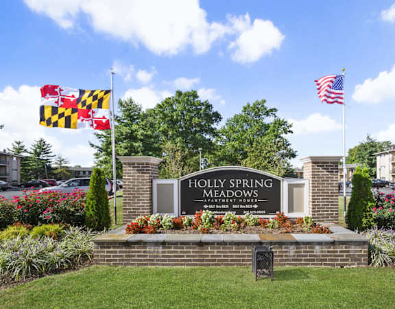 The entrance to Holly Spring Meadows is framed by a brick wall and adorned with a sign, a Maryland state flag, and an American flag.