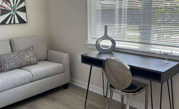 A room with a grey couch, a table with a chair and a dartboard on the wall.
