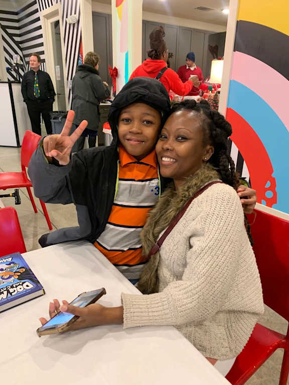 A young boy and a woman are sitting at a table with a book and a cell phone in front of them.