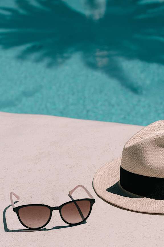 A pair of sunglasses and a hat are placed on a surface near a pool.