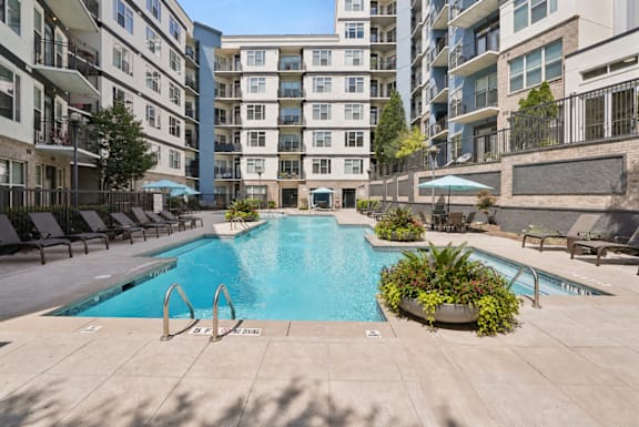 A pool surrounded by chairs and umbrellas in front of apartment buildings.