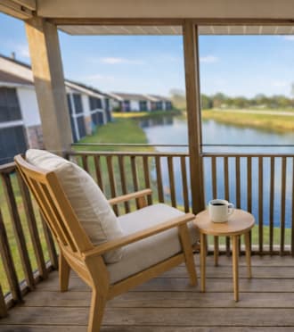 A wooden chair with a cushion is placed on a wooden balcony with a view of a lake and houses.