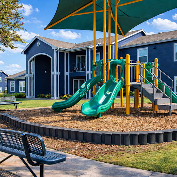 A playground with a green slide and a bench in front of apartment buildings.