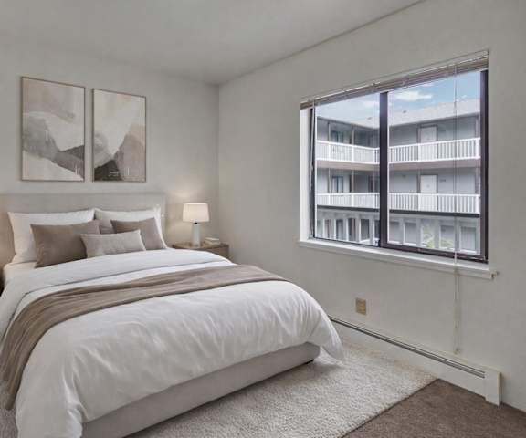 A view of the empty carpeted living room with a window, a fireplace, a door to the balcony, a rug, and the kitchen with white appliances and large, white cabinets at Silver Ridge Apartment Homes. 
