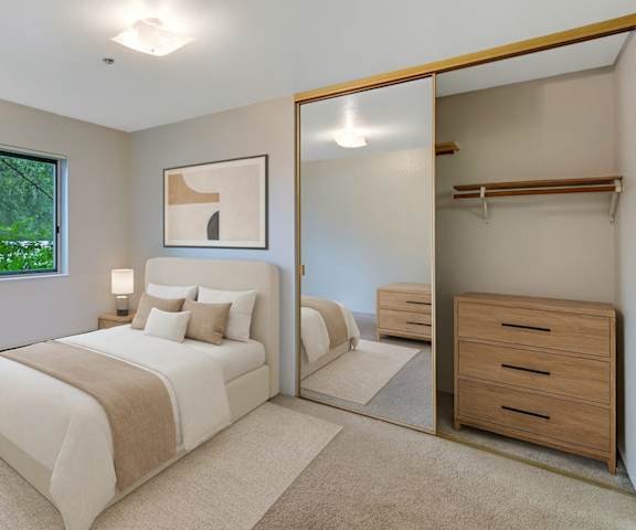 A large, empty carpeted bedroom with overhead lighting, a window showing the trees, and a closet at Susitna Ridge Apartment Homes. 