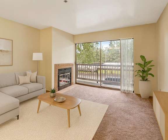A large, empty living room with carpeting, a fireplace, and a sliding glass door to the balcony at Susitna Ridge Apartment Homes. 