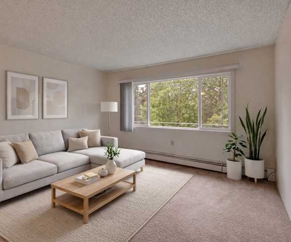 A view of the empty carpeted living room with a window and the kitchen with white appliances and wooden large cabinets at Taiga Apartment Homes. 